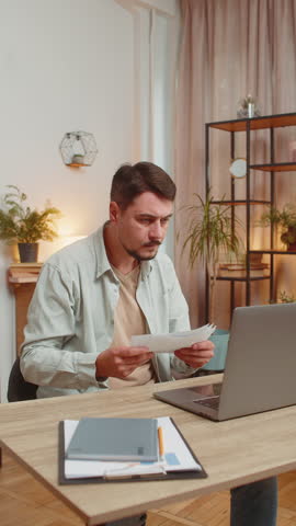 Stressed young man looking at unpaid bank debt bills, doing paperwork, planning budget calculate finances mortgage payments. Freelancer guy throws paper bills sitting at home office table. Vertical.