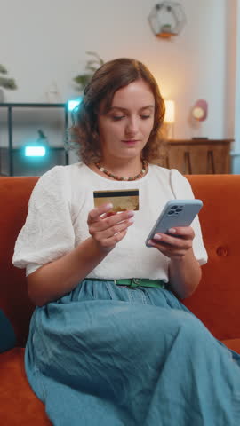 Happy young woman using credit bank card and smartphone while transferring money, purchases online shopping cashless order food delivery at home apartment indoors. Girl sitting on sofa couch. Vertical