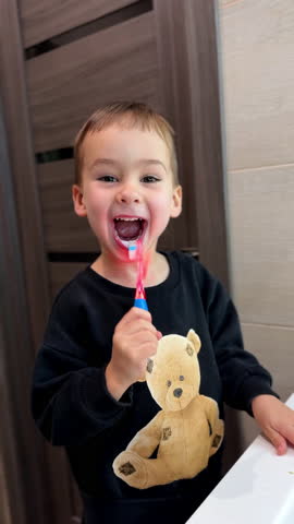 Baby boy wearing a sweater with teddy brushing teeth. Cheerful kid uses flashing tooth brush. Hygiene and health. Vertical video.
