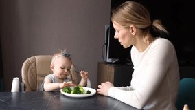 Young mother feeding baby girl broccoli at home. Toddler refuses healthy food. Concept of parenting, healthy eating, motherhood and nutrition for children. Caring mom with infant, natural light - Powered by Shutterstock - Get 15% off with code: PIKWIZARD15