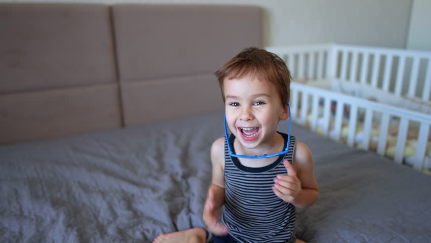 Adorable smiling Caucasian baby puts on his sunglasses. Active kid stands up and jumps on the big bed cheerfully.