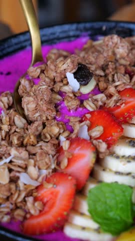 Vertical screen vibrant pitaya smoothie bowl garnished with granola, fruits, chia seeds, coconut flakes, mint, golden spoon lifting colorful creamy portion