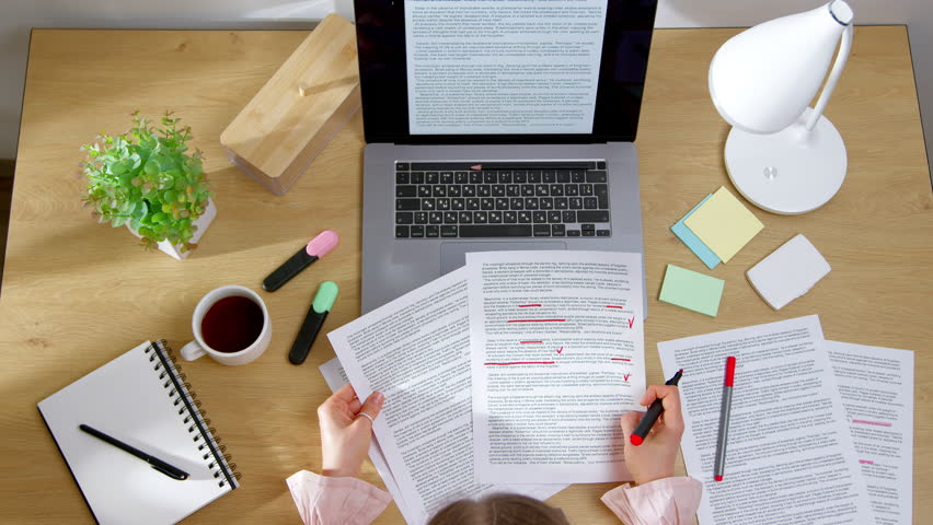 Top view of woman working on text for translation. Editor marks words in script on paper and laptop.	
