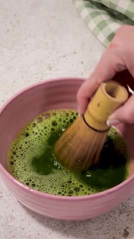 Woman preparing matcha tea using a bamboo whisk, creating a frothy beverage in a pink bowl