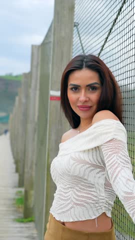 Female fashion model posing dynamically near metal fence, showcasing stylish outfit with confidence and grace during outdoor photoshoot in natural light