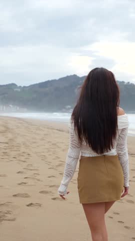 Brunette woman wearing casual attire standing on sandy beach, gazing serenely against cloudy sky with ocean backdrop, embodying carefree coastal lifestyle
