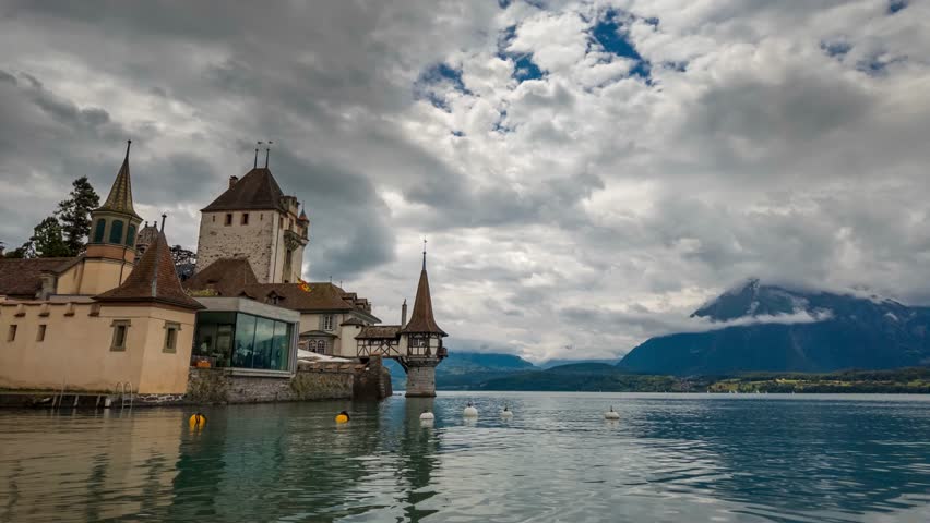 Timelapse of heavy clouds moving over Castle Oberhofen on the shore of Lake Thun in Switzerland, featuring medieval architecture with cloudy skies and Swiss Alps in the background. 4K UHD video.