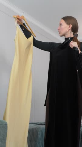 Young woman standing in bedroom, carefully comparing yellow dress and black jumpsuit hanging on hangers, contemplating fashion choice before deciding outfit for upcoming event vertical video