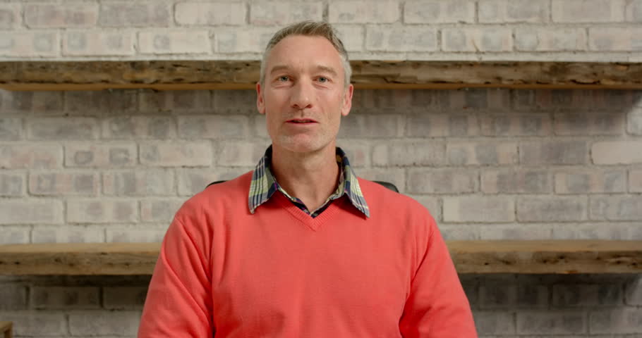 Close up of people talking to the camera, smiling man in casual sweater sitting in office with brick wall background. the person is fully visible, sitting, half body