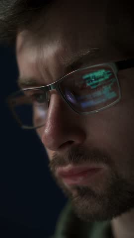 Close up of male software engineer in glasses working on big data, with code reflecting on his lenses. He is deeply engaged in programming and artificial intelligence tasks. Vertical video
