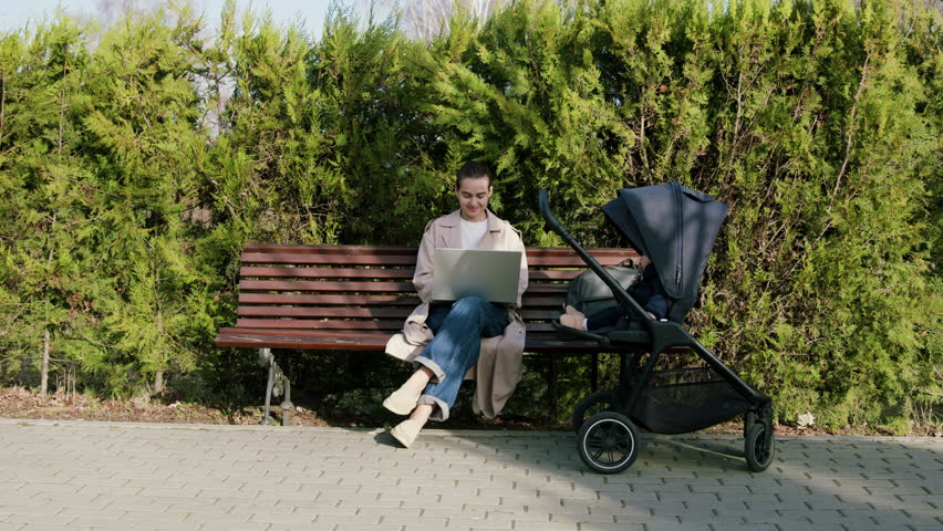 Young freelancer woman working from computer online remotely writing an email while baby is sitting in stroller and sleeping. Mother working in park outside while on maternity leave