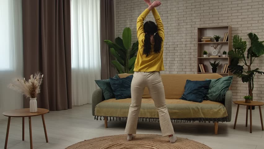 Woman in yellow top dancing twerk. The room is decorated with plants and has a cozy ambiance. Slow motion, back view