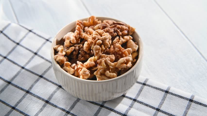 A woman's hand takes a peeled walnut kernel from a bowl filled with walnuts, preparing to enjoy a nutritious and delicious snack rich in protein and healthy fats