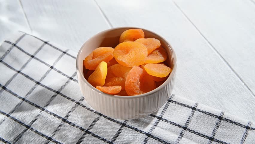 Woman's hand takes dried apricots from a bowl placed on a checkered napkin on a white wooden table, ready to enjoy a healthy and tasty snack