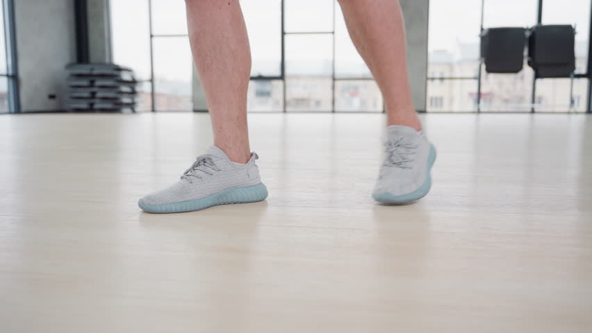 Male trainer leg view stretching both legs in side lunge on wooden gym floor with sneakers showcasing muscle tone under large windows and bright studio lighting in modern fitness environment