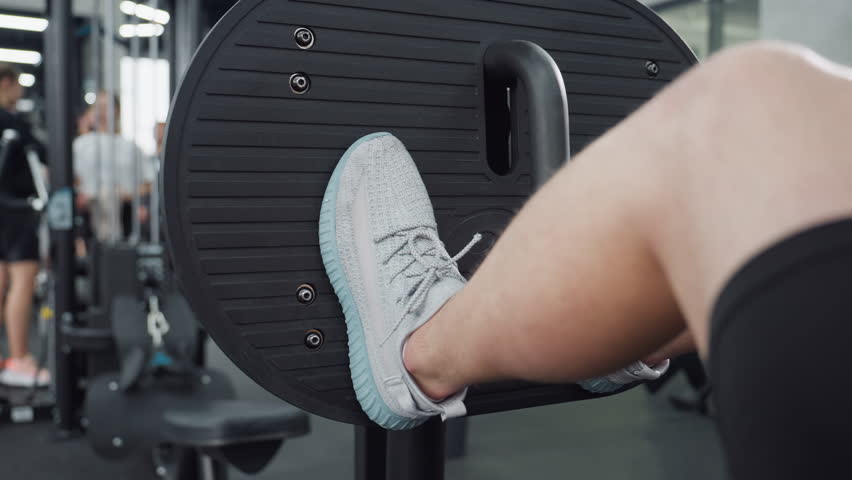 Man works out on leg press machine, driving foot plate to engage quadriceps and glutes, wearing sneakers, positioned under bright modern gym lighting with other athletes blurred in background