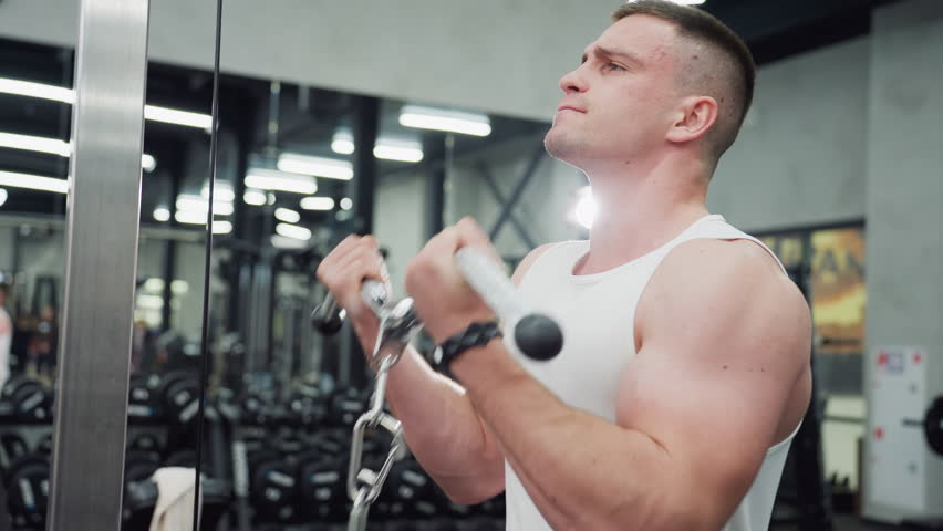 Athlete grips cable machine handle in modern gym and powers through intense arm curl set, brow furrowed, muscles flexing, reflecting disciplined strength training routine in fitness center