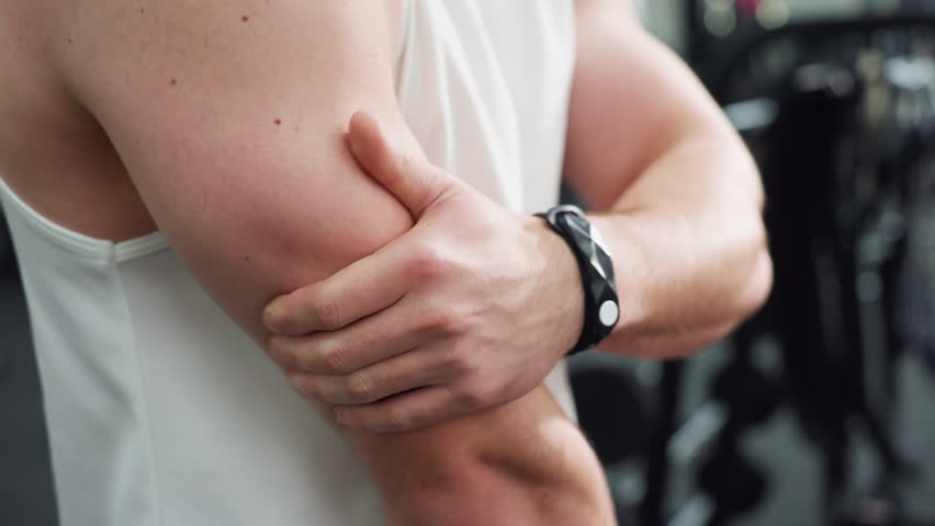 Body builder slaps hands against flexed arm muscles to loosen tension post workout, white singlet and black shorts visible, gym equipment blurred in background, focus on muscular definition