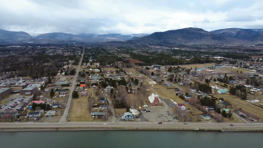 Panoramic aerial view of a village located between the sea and the mountains in a dull, winter-yellowed setting under a gray, cloudy sky. Maria, Quebec, Canada, 2025.