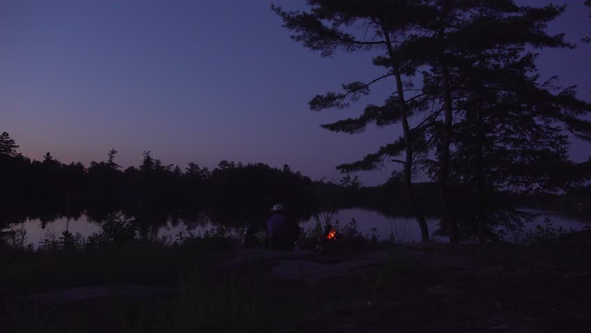 A man wearing a Tilley hat sits beside a campfire on the shoreline of a reflective lake at dusk.