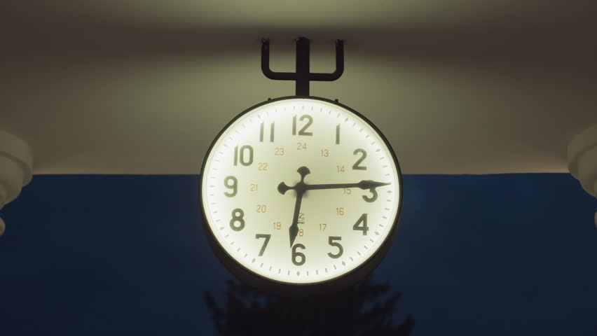 Close up of glowing round clock hanging on metal fork-like bracket under roof structure, surrounded by darkness of evening sky, casting soft ambient light over architectural elements of building top