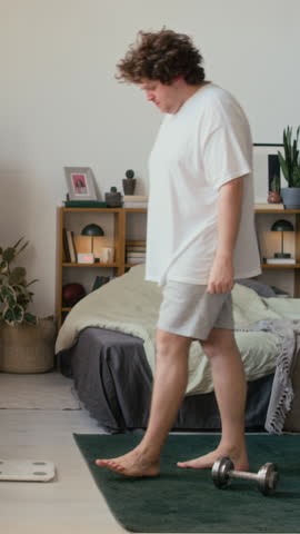 Vertical shot of young adult man wearing comfy outfit stepping on white scale to check his body weight