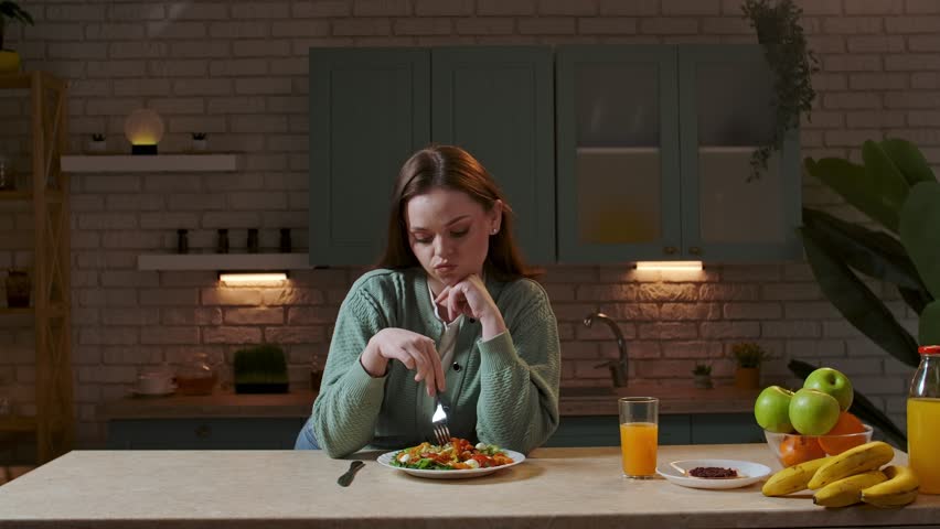 Healthy eating concept. Woman at the table in the kitchen preparing healthy vegetable salad for dinner, woman mixing salad with fork on the plate, no appetite, sad unhappy expression. Slow motion