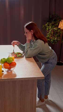 Healthy eating concept. Woman at the table in the kitchen preparing healthy vegetable salad for dinner, woman mixing salad with fork on the plate, no appetite, sad unhappy expression. Vertical video