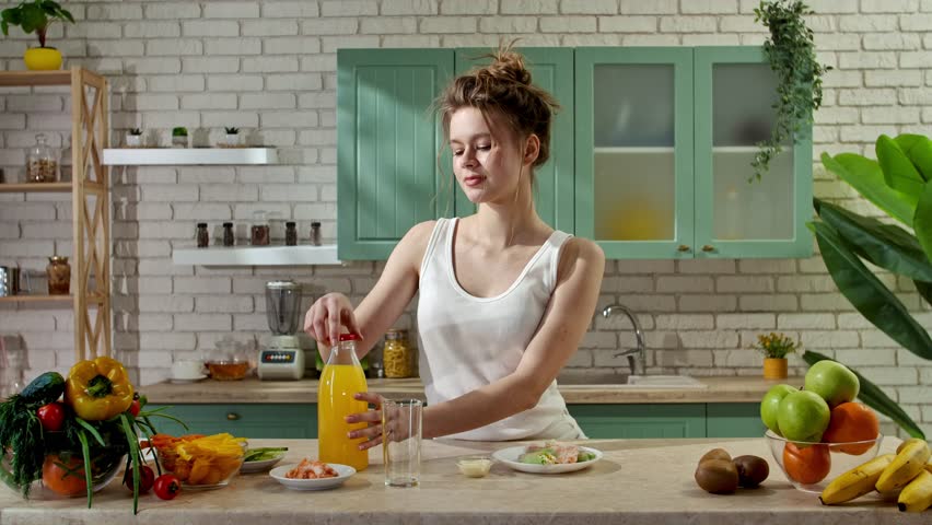 Healthy eating concept. Young woman at the table in the kitchen preparing spring rolls for healthy lunch, pours orange juice in glass, eats fresh spring rolls, and drinks juice. Slow motion.