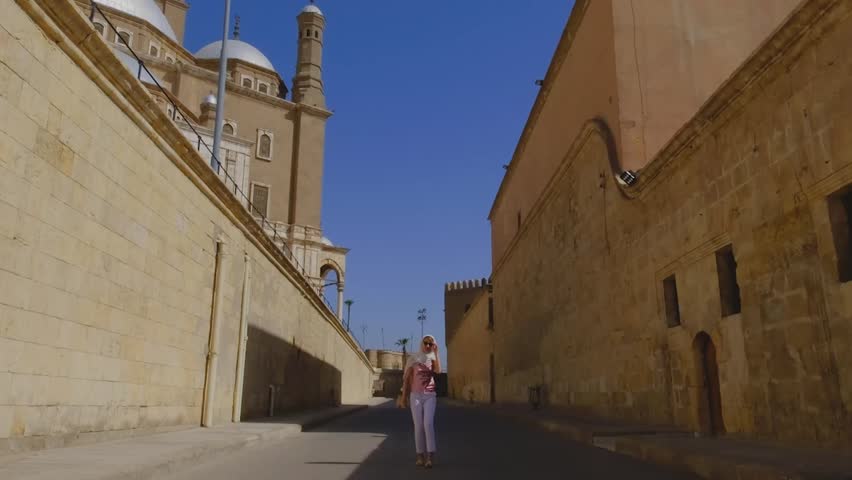A woman walks down a historic street in Cairo, Egypt, with the grand mosque in the background. 