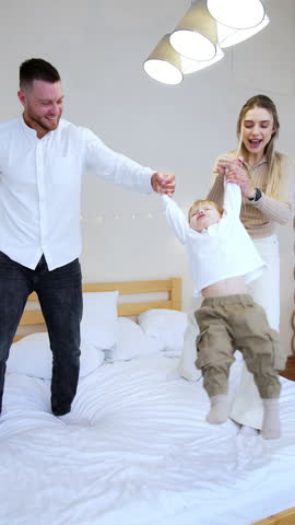 Caucasian parents waving their baby by the hands. Mom and dad play with their child on the bed. Happy family together. Vertical video.