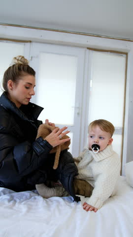 Woman wearing black jacket sits on the bed. Mother dresses her baby boy to go for a walk. Vertical video.