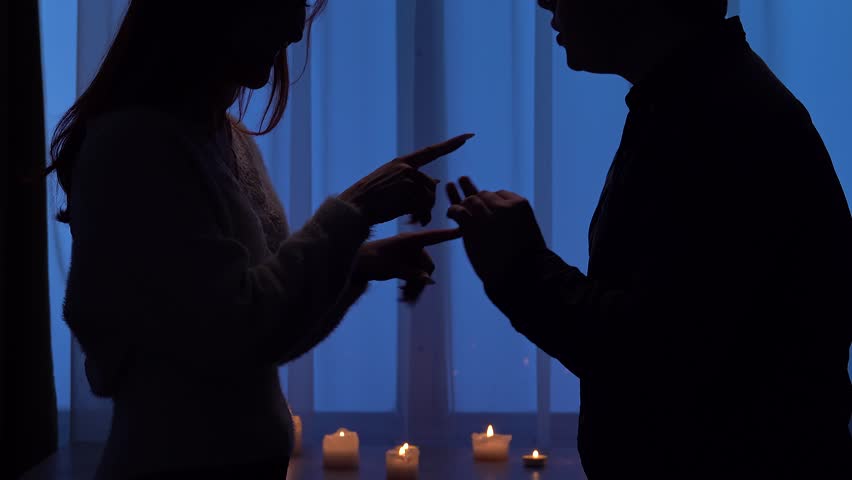 Silhouettes of a man and a woman facing each other in a dimly lit room and arguing, against a background of blue curtains and lit candles. Relationship Concept.
