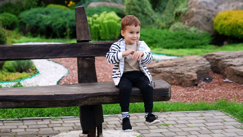 Smiling Caucasian toddler in stripy blazer sits on the bench in the park. Lovely kid waves his hands playfully.