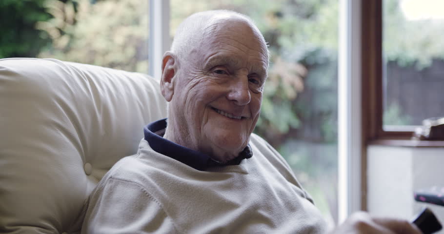 Face, old man and watching television to relax, smile and in nursing home for retirement, happiness and peace. Portrait, senior citizen and mature male with remote for tv, calm and joyful in lounge