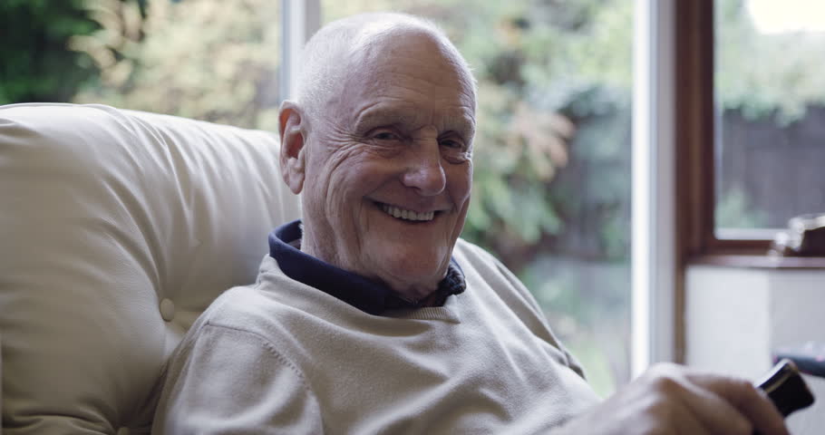 Face, old man and watching television to relax, smile and in nursing home for retirement, happiness and peace. Portrait, senior citizen and mature male with remote for tv, calm and joyful in lounge