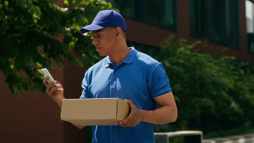 Small business concept African American courier man searching address with mobile phone app walking in city delivery guy male post service e-commerce shopping order hold carrying box parcel difficult