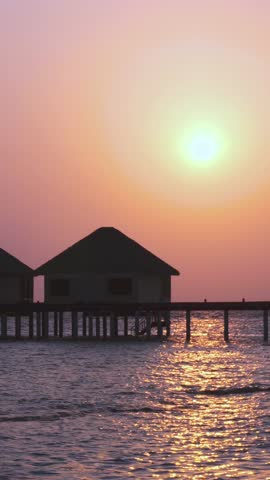 Sunset scene at Maldives seashore with luxury water lodges through palm tree leaves. Travel destinations. Vertical footage