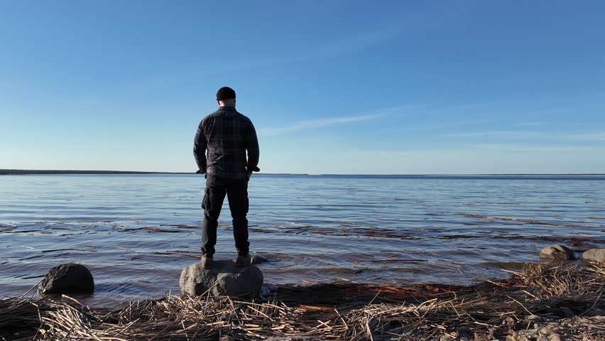 A man stands on a rock on the beach and looks at the landscape. view from the back.