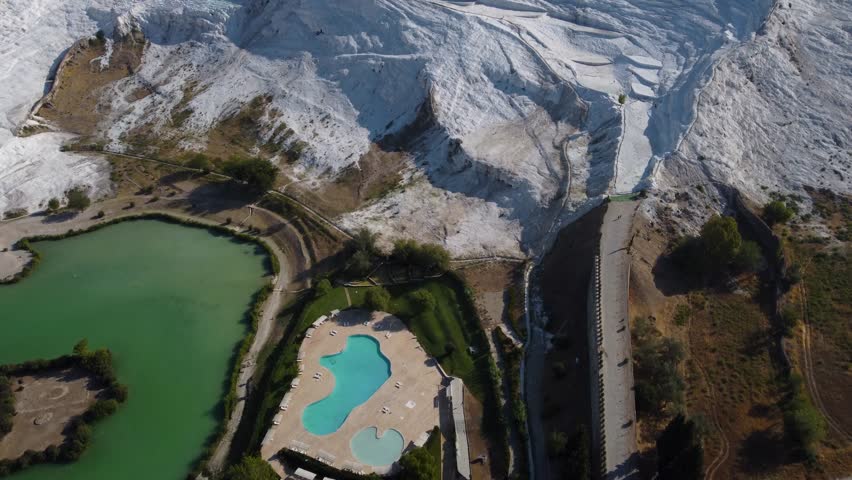 Forward tilt-up drone glides over Pamukkale’s white travertine pools and terraces, then rises to reveal the yellow-brown plateau and the ancient ruins of Hierapolis.