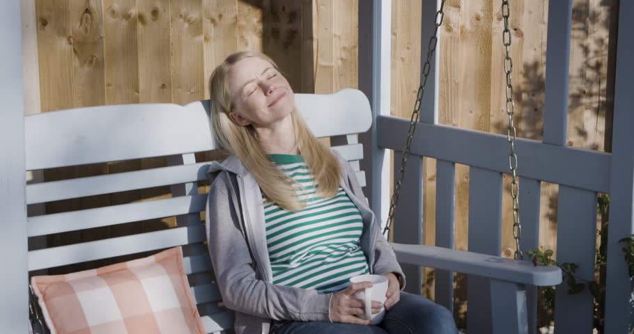 Woman, relax and happy in sunshine, outdoor fresh air and peaceful break for mental health, wellness and coffee. Summer, happiness and smile with female relaxing on bench, backyard and calm freedom
