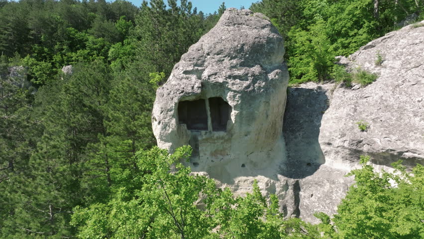 Stone-carved cave structure with square window cut into cliffside rock, positioned above forested slopes and rugged landscape in remote countryside.