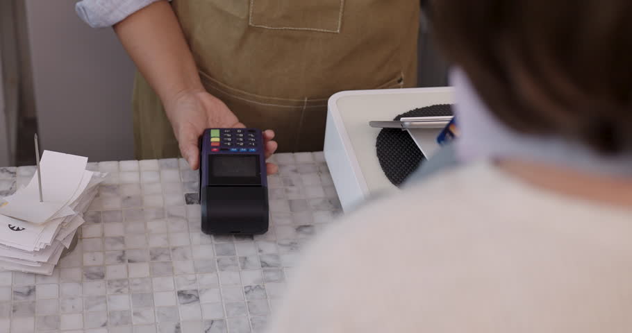 Female barista in brown apron holding credit card terminal while customer hand inserts blue card during payment process in cafe, motion indicates quick transaction and efficiency