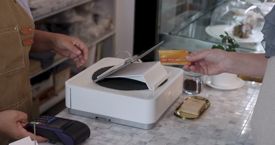 Customer hand moving forward with gold credit card toward wireless terminal as barista holds machine at counter inside cafe, seamless contactless payment action in progress