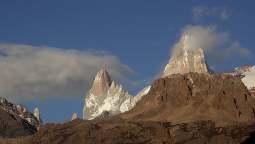 Clouds swirling around snow-capped peaks of Mount Fitz Roy and Poincenot, with rocky foreground under blue sky in El Chalten, Patagonia, Argentina