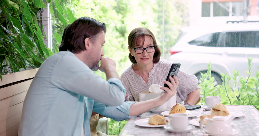 Older man showing something on smartphone to woman at cafe, fingers point to screen while she listens with curiosity, coffee and pastries on table nearby