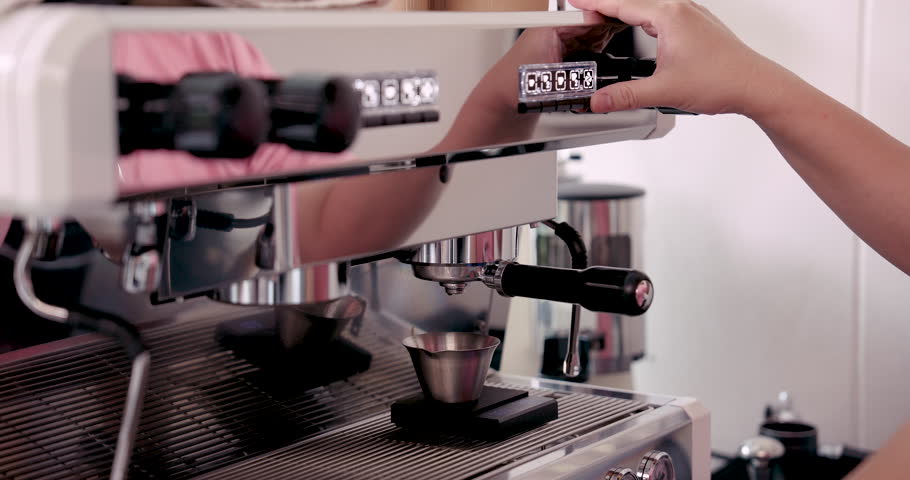 Cafe barista activating professional espresso machine with finger to begin brewing coffee, brown liquid flows steadily into metal cup, demonstrating fresh preparation