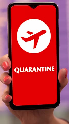 Close up, woman, in protective mask, holds smartphone with airplane icon, quarantine sign, on screen, in front of flight information board in departures terminal, at airport.