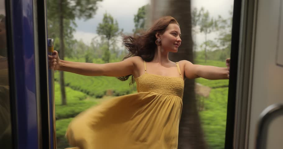 A woman in a yellow dress leans out of a train door, embracing the scenic beauty of Sri Lanka's tea plantations. Ideal for travel, adventure, and lifestyle promotions showcasing extreme attractions