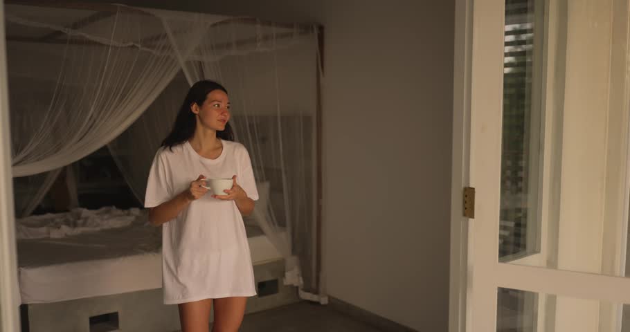 A young woman in a loose white shirt enjoys a warm cup of coffee in a serene tropical villa. Standing near a bright doorway, she embraces the morning light and tranquil atmosphere.
